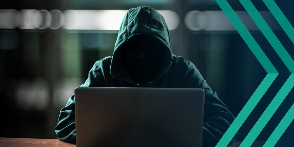 Image of hooded figure in front of laptop, representing article reporting the rising cybersecurity risk of recruitment fraud and job scams