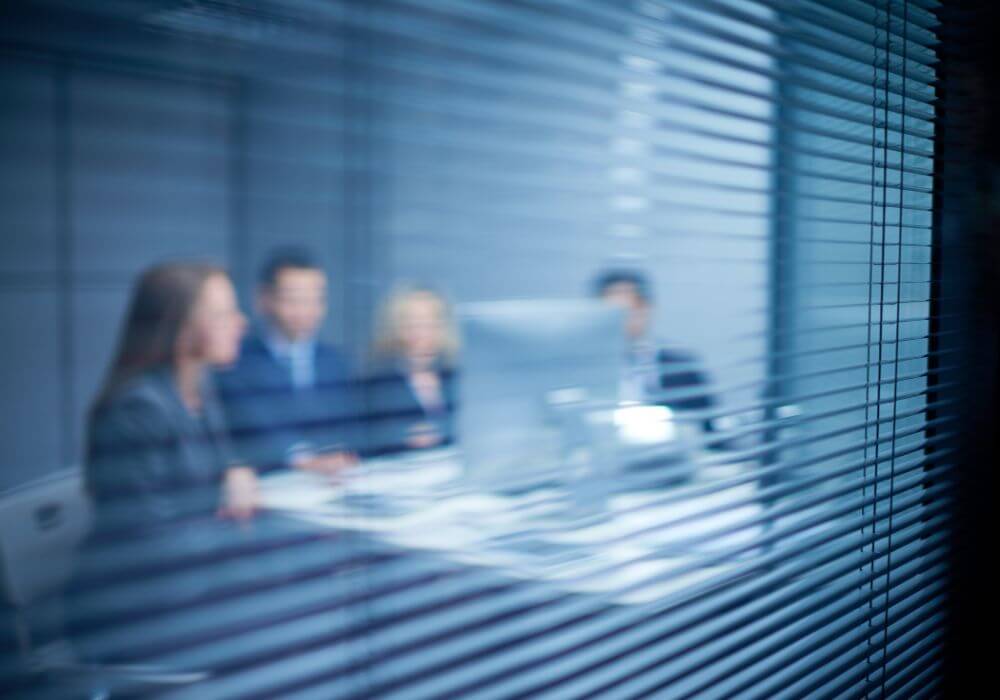 Photo of office meeting behind blinds, implying hidden risks for recruitment of contractor liability