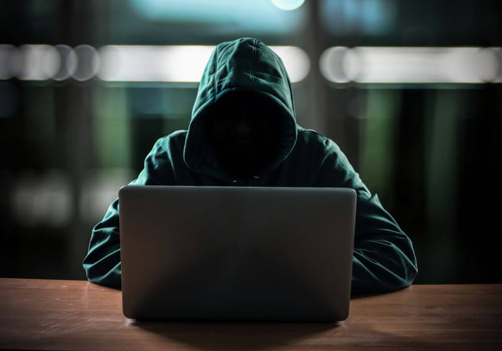 Image of hooded figure in front of laptop, representing article reporting the rising cybersecurity risk of recruitment fraud and job scams